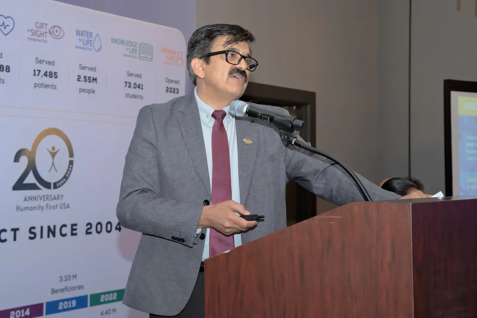 A speaker addresses the Gift of Health Gala from the podium beside the Humanity First USA 20th anniversary display