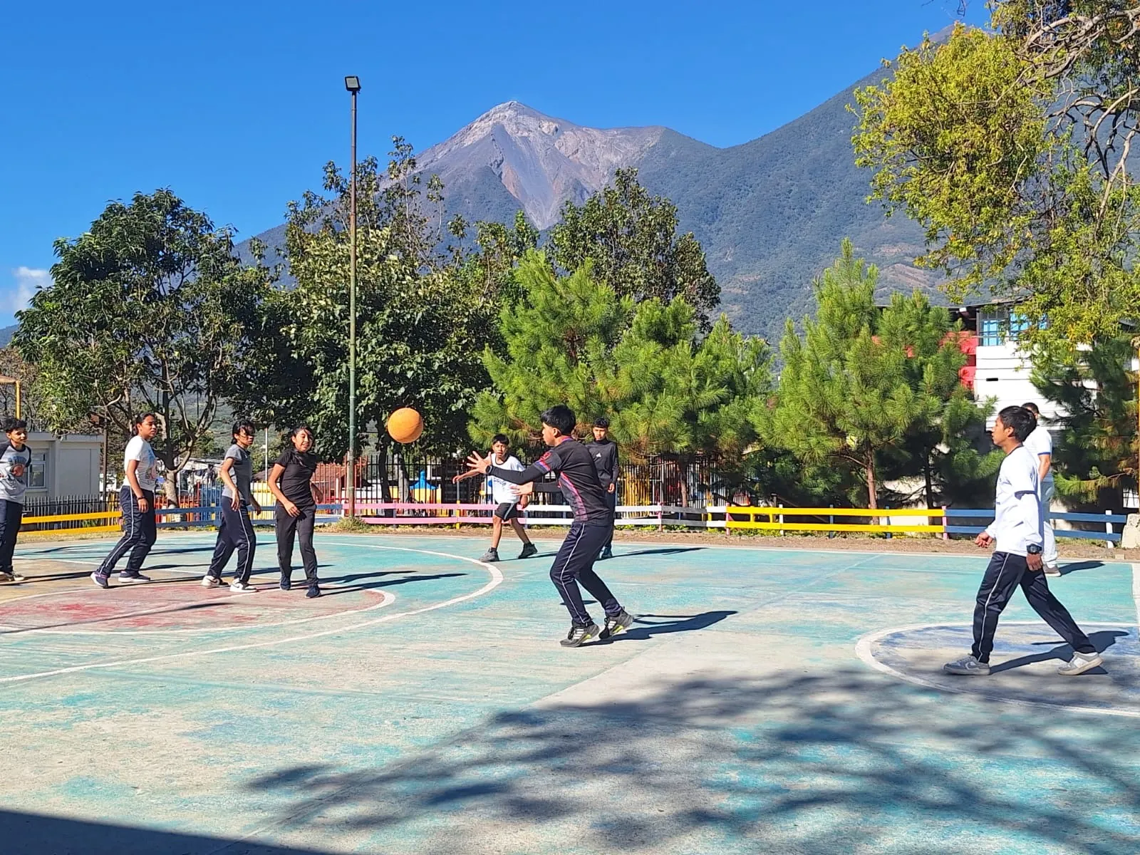 Masroor School students play basketball under a clear sky with a volcano beyond the trees