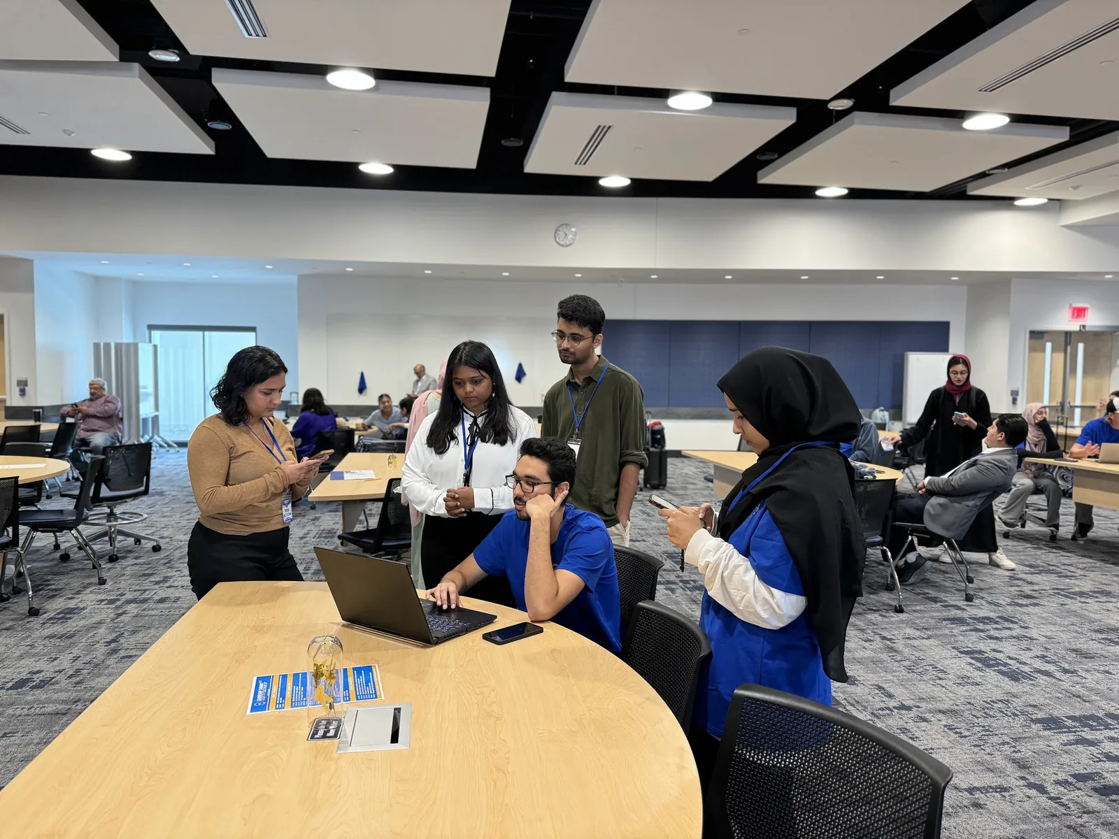 A team of students gathers around a laptop at a workshop table, one seated at the keyboard while the others follow along and check their phones, at the HFSD Leadership Summit 2024