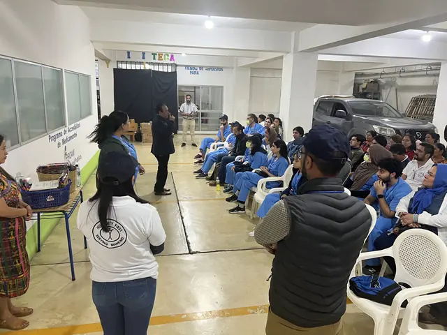 Students in blue scrubs and community members gather along the walls of a clinic hall for an opening program in Guatemala.