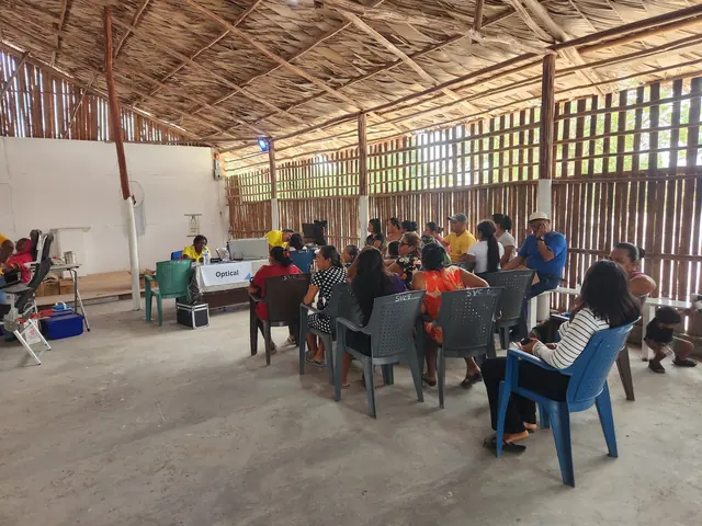 Patients sit in plastic chairs in a quiet corner of the open air clinic in Guyana as volunteers staff a table nearby.