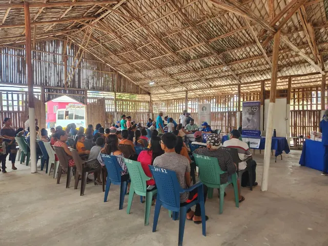 Community members sit in rows of colorful plastic chairs facing the front of the thatched roof clinic pavilion in Guyana.