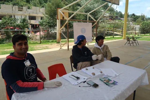 A smiling volunteer in gloves sits at a registration table under a covered court as patients wait nearby at the jornada médica in Mexico.