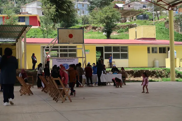 Community members line up at registration tables under a covered court beside a yellow school building during the jornada médica in Mexico.
