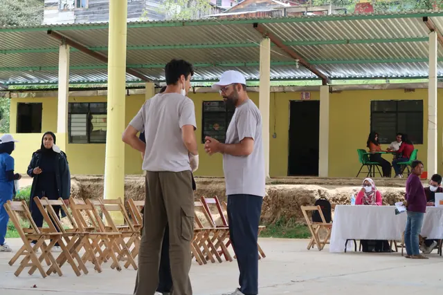 Two masked student volunteers talk beside stacks of folding chairs as a colleague works at a registration table in the clinic in Mexico.