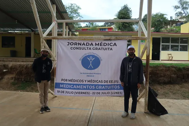 Two volunteers stand on either side of a Jornada Medica banner announcing free consultations and medicines at the clinic site in Mexico.