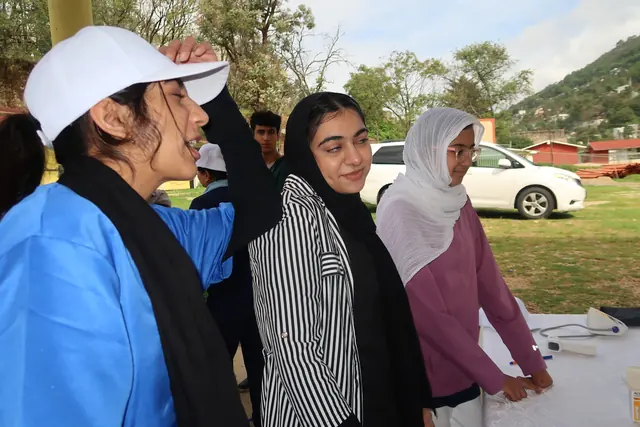 Three students in scrubs, a white cap, and headscarves smile and talk at a clinic station under the open sky in Mexico.