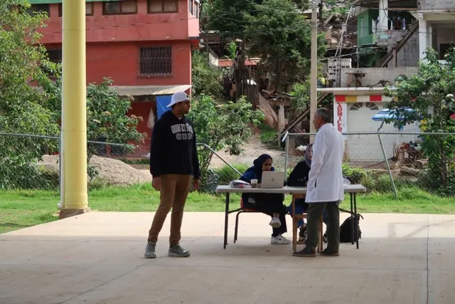 A physician in a white coat stands at a consultation table under a covered court as volunteers and patients gather at the clinic in Mexico.