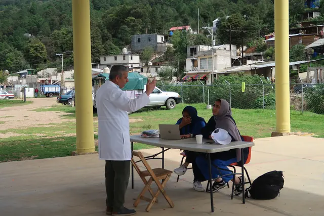 A physician in a white coat speaks with two students working on a laptop under the covered court of the clinic in Mexico.