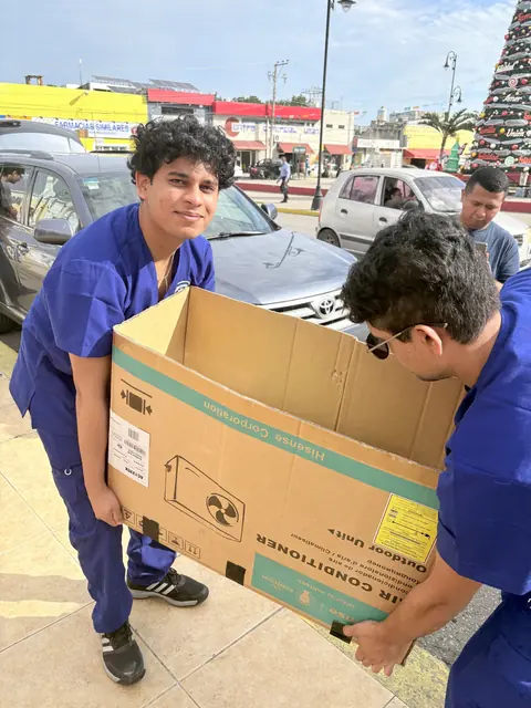Two smiling volunteers in blue scrubs carry a large cardboard box of supplies across the street at the Gift of Health mission in Mexico.