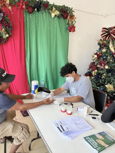 A masked volunteer checks a patient's blood sugar at a table beside a decorated holiday tree at the Gift of Health mission in Mexico.