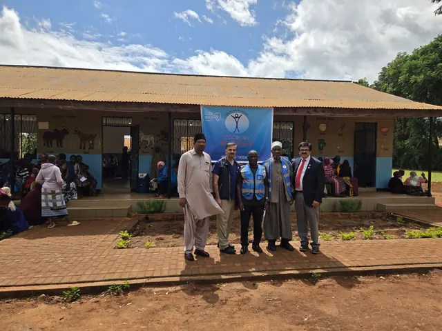 Mission leaders in Humanity First vests stand before a Gift of Health banner at the school clinic as patients wait on the veranda in Tanzania.