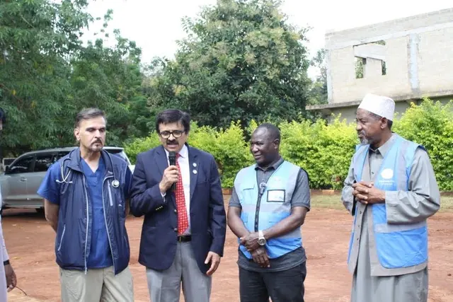 Mission leaders in Humanity First vests and suits address the community outdoors in front of green trees at the Tanzania clinic.