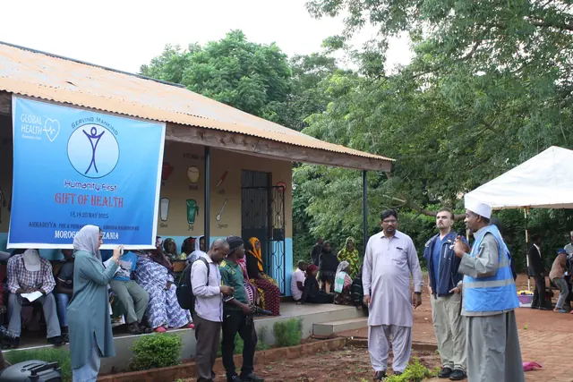 Volunteers and patients gather outside the clinic building beneath a Gift of Health banner with a white tent nearby in Tanzania.