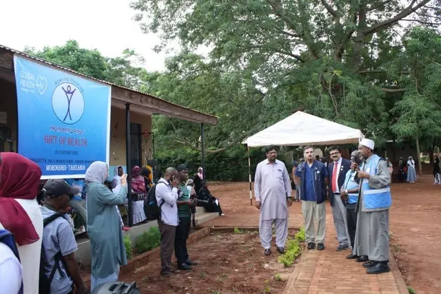 Mission leaders and volunteers talk outside the clinic beside a Humanity First banner and white tent as patients wait in Tanzania.