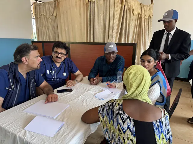 Physicians in navy scrubs, an interpreter, and a student in a Humanity First vest consult with a patient at a table in Tanzania.