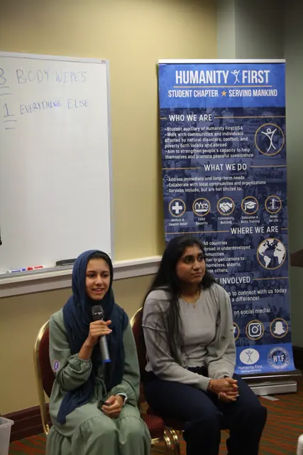 Two panelists seated before a Humanity First banner share their experiences with a microphone at the HFSD Leadership Summit '23.