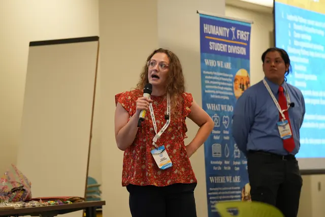 A presenter speaks into a microphone beside a Humanity First Student Division banner as a colleague looks on at the HFSD Leadership Summit 2026.