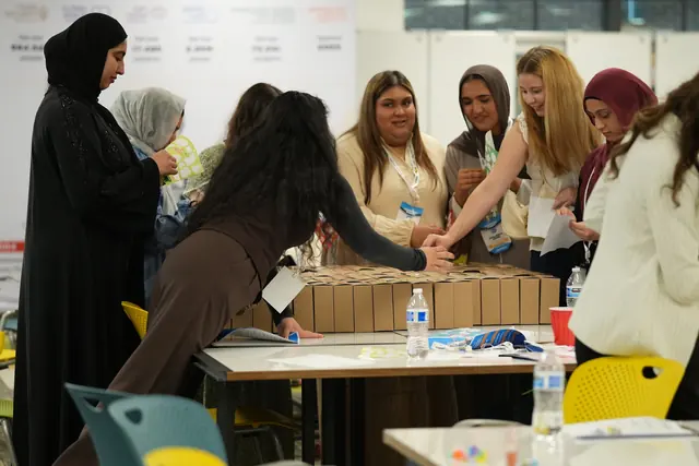 A group of students reaches across a table to add items to rows of finished kits at the HFSD Leadership Summit 2026.