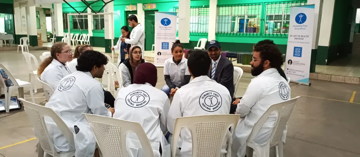 Volunteers in white Humanity First coats seated in a circle of plastic chairs, with a Humanity First Gift of Health Program banner in the background.